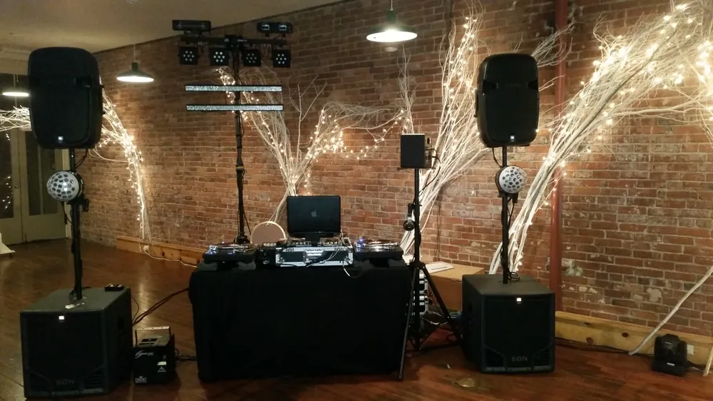 The rig with a black-draped table against a brick wall: speakers on stands, light bars and white branches with fairy lights